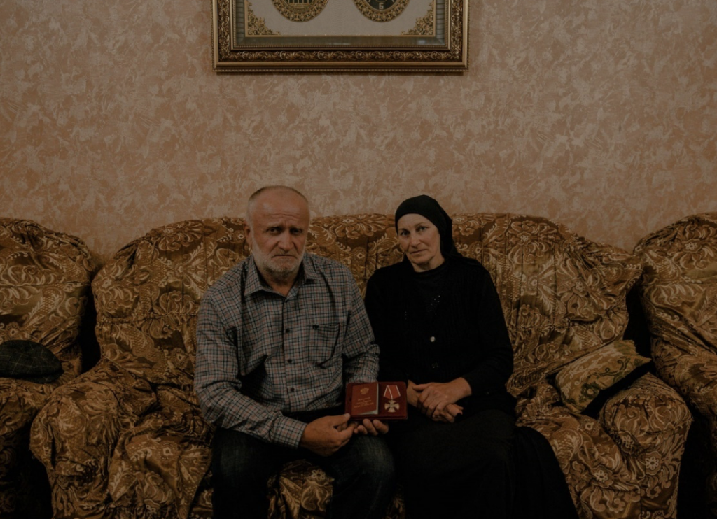 A man and woman sit on a patterned couch holding an Order of Courage medal that was given to them on behalf of their son...