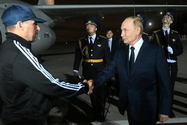 A man in casual clothing and a cap shakes hands with Vladimir Putin. A plane and an honor guard are in the background. 