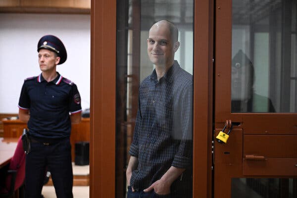 Evan Gershkovich in a glass defendant’s box smiles at the camera.